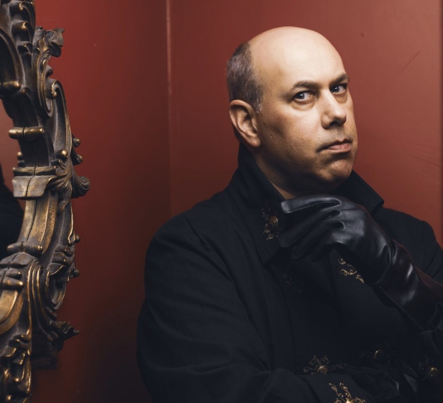 Jason Carl in einem schwarzen Anzug mit schwarzen Handschuhen vor einer matt roten Wand mit einem Spiegel zu seiner rechten Seite.